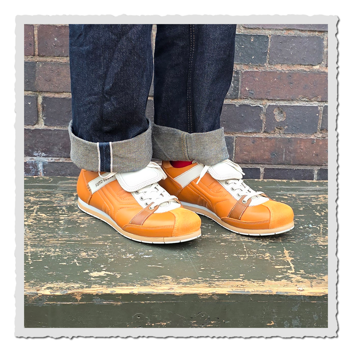 Orange Schnürschuhe mit heller Lasche an einer Person mit dunklen Jeans vor Backsteinwand auf grünem Holzpodest.